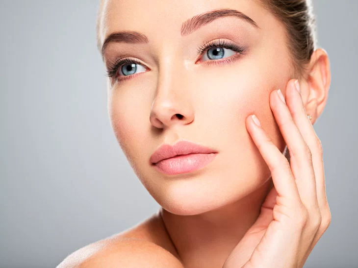 a imagem mostra uma mulher branca com olhos azuis e lábios rosados. Ela está parada na frente de uma parede cinza, com a uma das mãos no rosto.