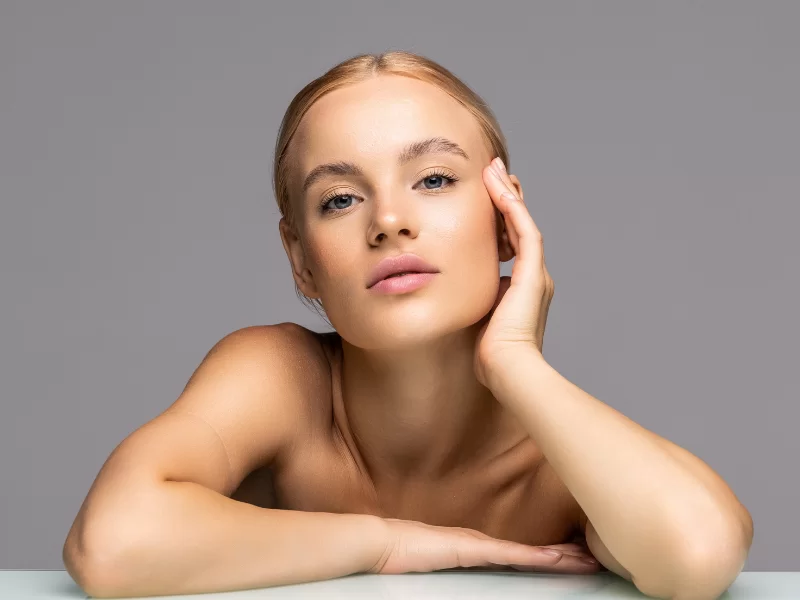 A imagem mostra uma mulher branca, com cabelos lisos e loiros, presos em um coque para trás. Ela está olhando para frente, com o braço direito recostado sobre uma superfície branca e a mão esquerda tocando a lateral do rosto. O fundo da imagem é cinza.