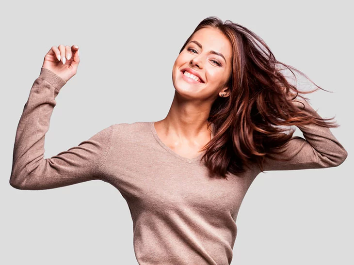 A imagem, de fundo branco, mostra uma mulher jovem, de pele clara e cabelos castanhos pulando. Ela está sorrindo para a câmera e veste uma blusa de manga longa da cor bege.