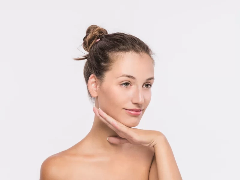 A imagem mostra uma mulher, com cabelos presos em um coque e com a mão esquerda apoiando o queixo. O fundo da imagem é cinza.