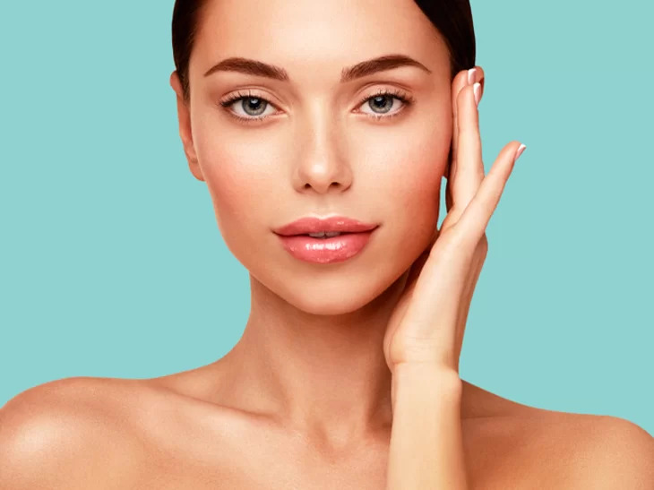 Na imagem aparece uma mulher branca, com cabelos pretos, para em frente a uma parede azul esverdeada. Ela está com uma das mãos na lateral do rosto.