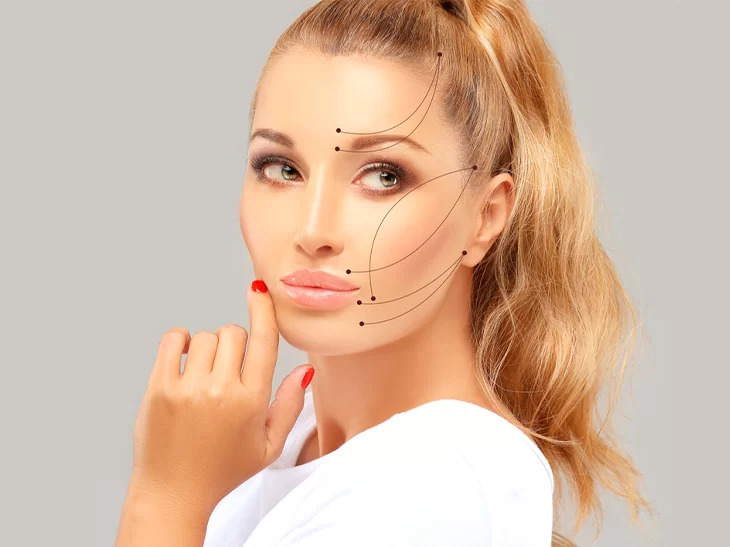 Na imagem tem uma mulher branca e loira, com os cabelos presos em um rabo de cavalo, usando roupa branca. Ela está com as unhas vermelhas e uma das mãos próxima do rosto. É possível ver alguns traços em seu rosto, destacando detalhes melhorados através dos procedimentos estéticos.