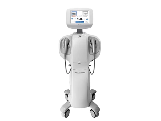 na imagem, é possível ver um aparelho branco e cinza, com quatro rodas, um painel touch screen e dois dispositivos utilizados para realizar o procedimento.