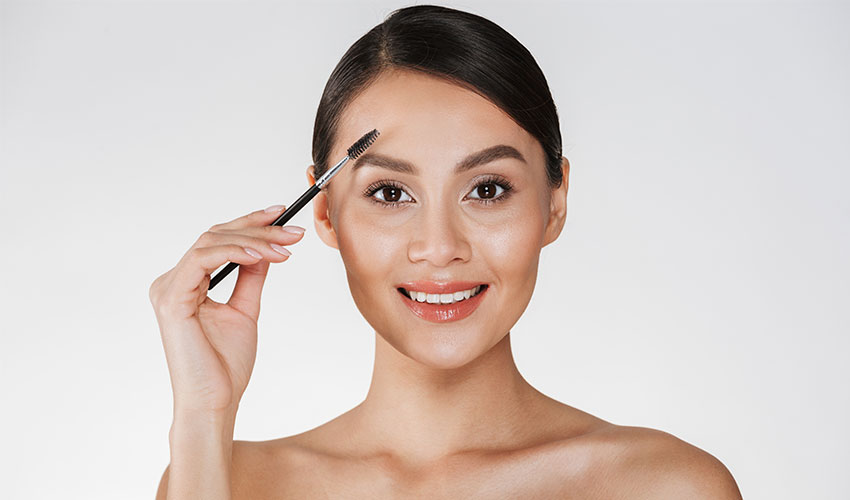 A imagem mostra o rosto de uma mulher jovem, de pele clara e cabelos pretos amarrados em um coque. Ela olha para a câmera penteia a sobrancelha esquerda com um pente de sobrancelhas.