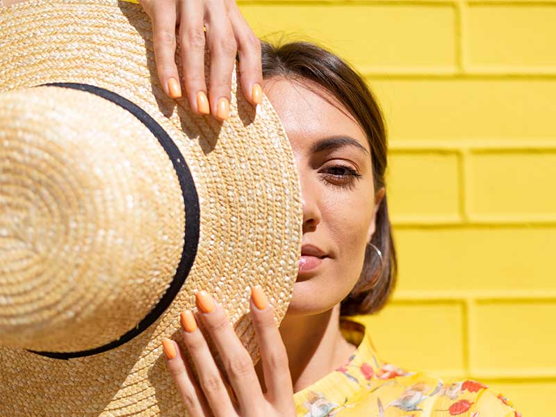 A imagem mostra uma mulher, de cabelos castanhos e curtos, segurando um chapéu de praia na frente da metade esquerda do rosto. Sua pele é branca e atrás dela é possível ver uma parede de tijolos amarelos.