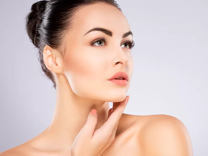 a imagem mostra uma mulher branca com cabelos pretos presos em um coque. Seu maxilar está marcado e ela passa uma de suas mãos sob o queixo. Atrás dela há uma parede cinza claro.