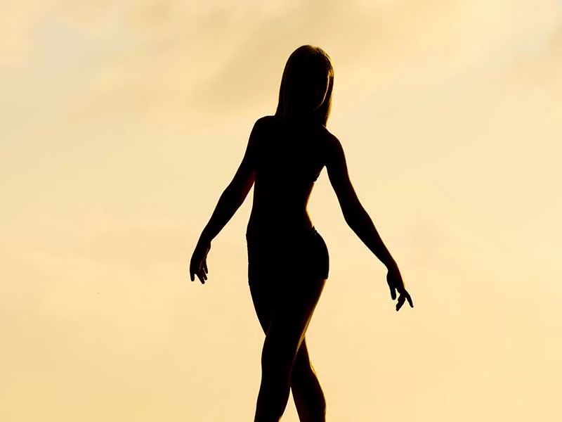 O fundo da imagem é colorido por um laranja clarinho e esfumado e, sobre ele, é possível ver a silhueta toda escura de uma mulher, na ponta dos pés e com os braços posicionados graciosamente ao lado do corpo.