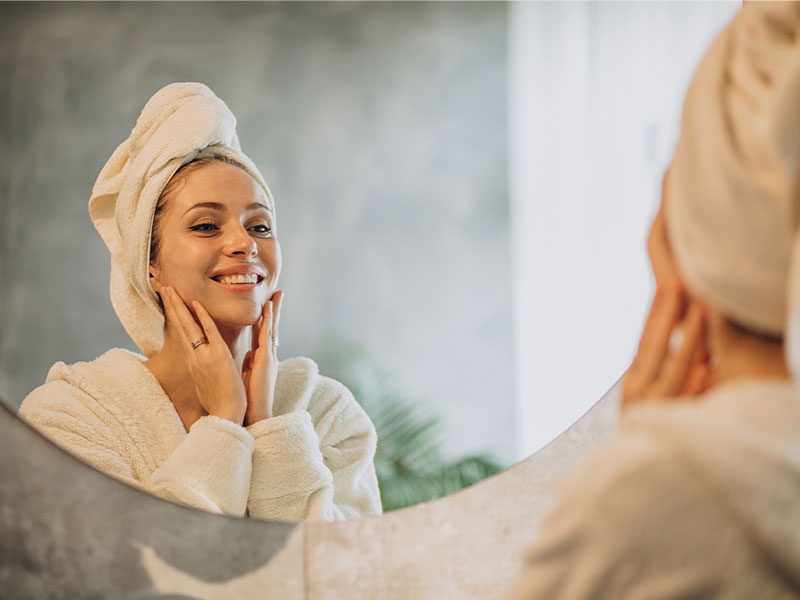 A imagem mostra uma mulher branca sorrindo após o banho, com uma toalha branca enrolada na cabeça e suas mãos abaixo do rosto. Ela está se olhando no espelho e vestindo um robe branco. O espelho é redondo e está em uma parede cinza, no reflexo é possível ver algumas plantas verdes e uma parede cinza.