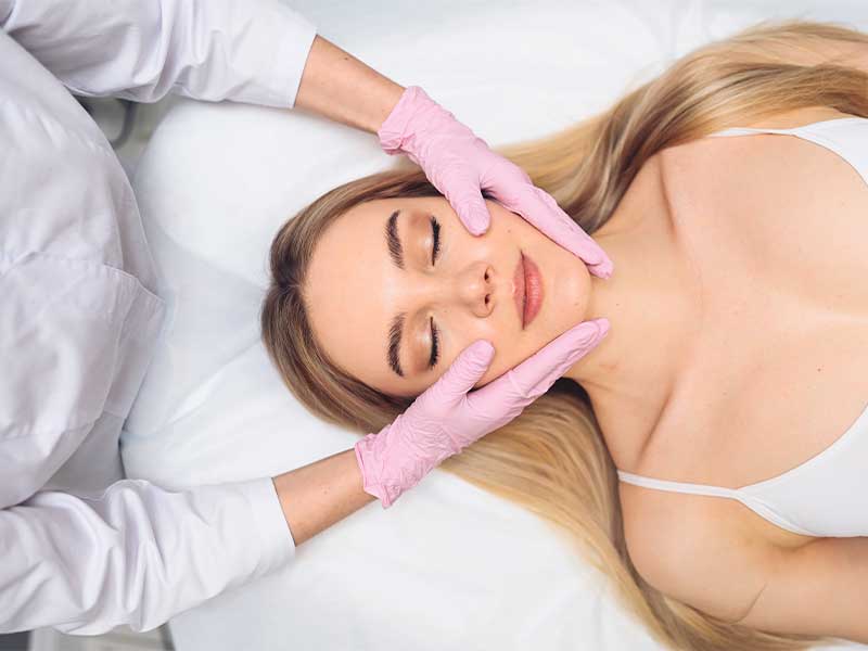 A imagem mostra uma mulher loira deitada em uma maca branca. Ela está de olhos fechados, vestindo uma regata branca. Na imagem também há uma esteticista com luvas rosas e jaleco branco. Ela está com as mãos no rosto da paciente, que está relaxada.