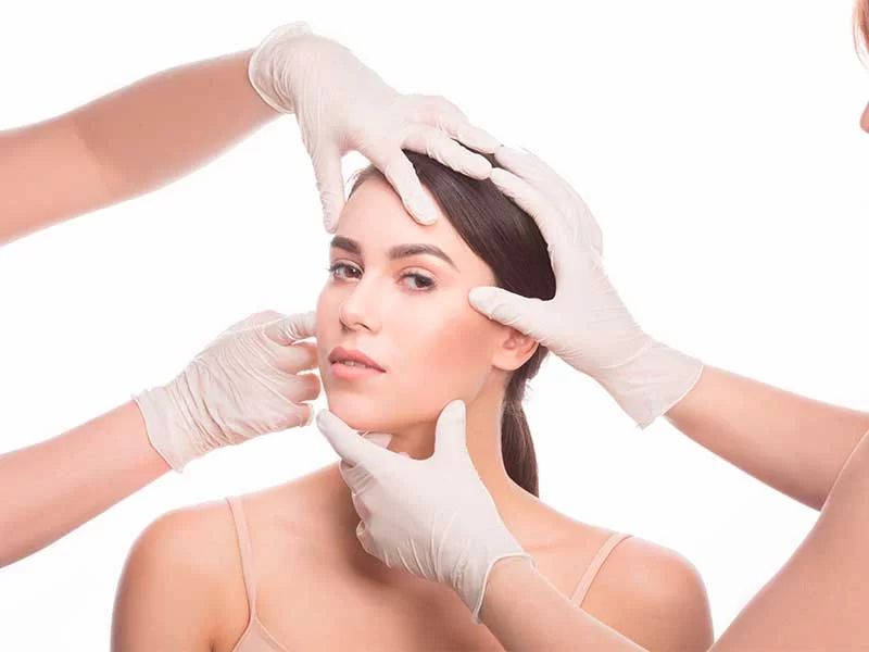 Na imagem há uma mulher com cabelos castanhos presos em um rabo de cavalo. Ao redor dela há dois pares de mãos com luvas cirúrgicas analisando seu rosto.