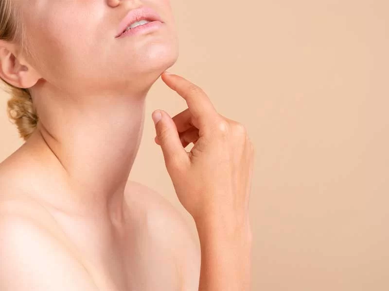 Na imagem há a parte do corpo de uma mulher branca, a região do colo e pescoço, com parte dos lábios aparecendo. Ela está com o dedo indicador tocando seu queixo, enquanto o levanta para mostrar mais o pescoço.