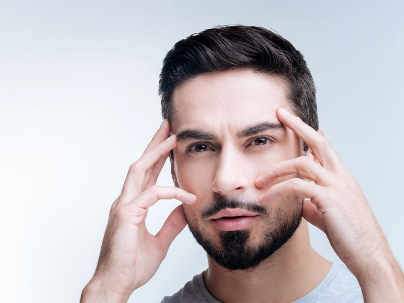 Na imagem, temos um homem com cabelos escuros e alguns fios brancos. Ele possui barba e olhos escuros também. Ele está usando uma roupa cinza e está com as mãos perto dos olhos, destacando o olhar.