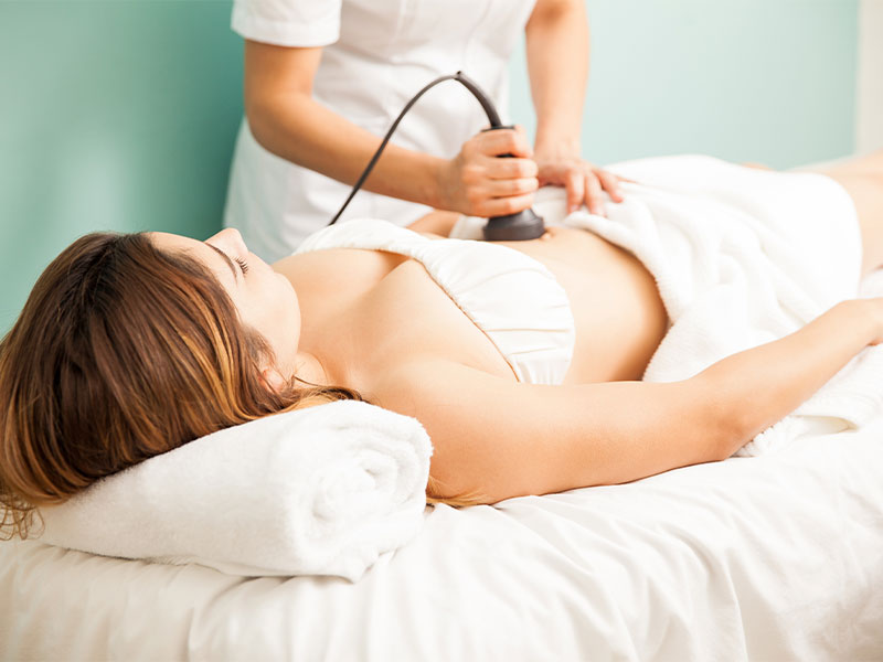 A imagem mostra uma mulher deitada em uma maca, enquanto a profissional está aplicando uma tecnologia em seu abdômen para tratar gordura localizada. Ambas as mulheres usam peças de roupas brancas, mesma cor que há nas toalhas.